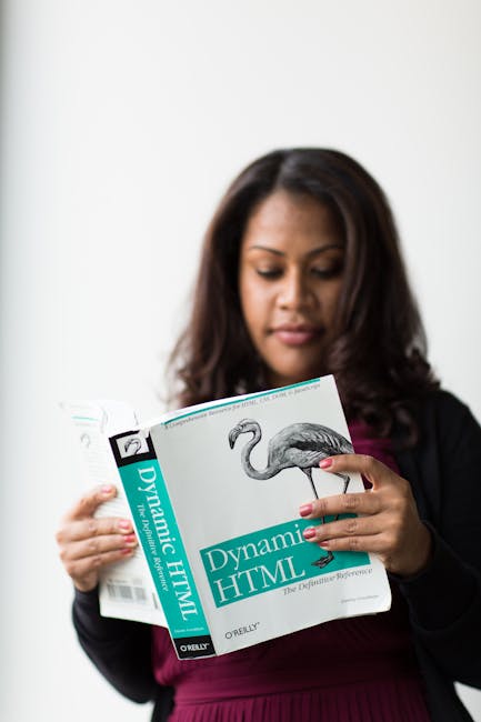 A woman with medium-length dark hair is holding and reading a book titled 'Dynamic HTML' by O'Reilly, featuring a black-and-white illustration of a flamingo on the cover. She is dressed in a black bla